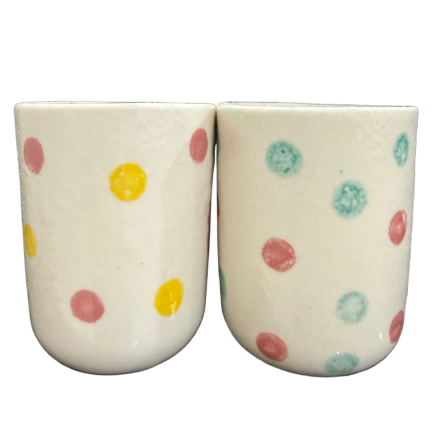 Two cream-colored ceramic cups with colorful hand-painted polka dots in yellow, pink, and teal, set against a white background.